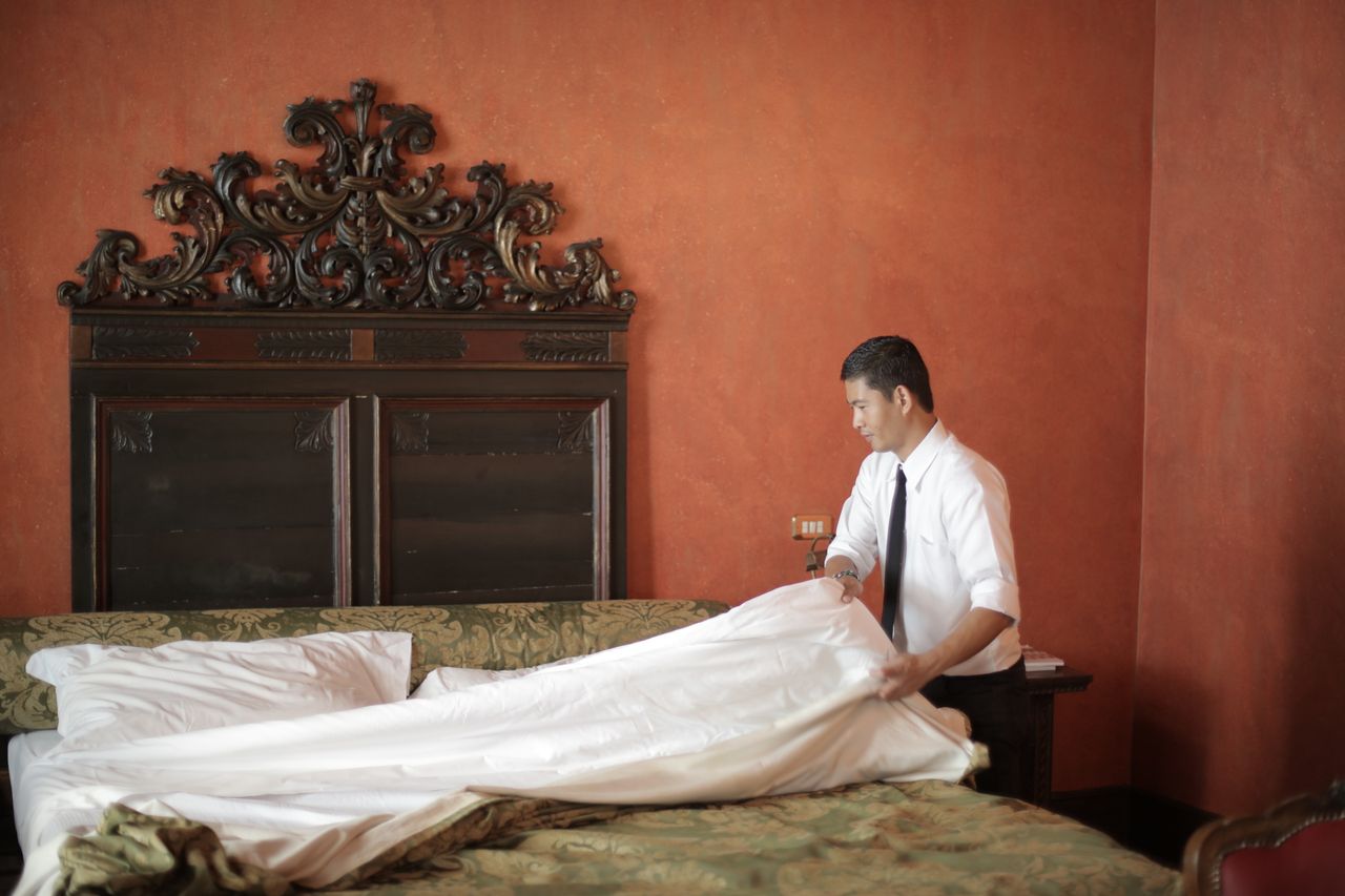services-04 A hotel staff member in formal attire making a bed in a luxurious bedroom setting.